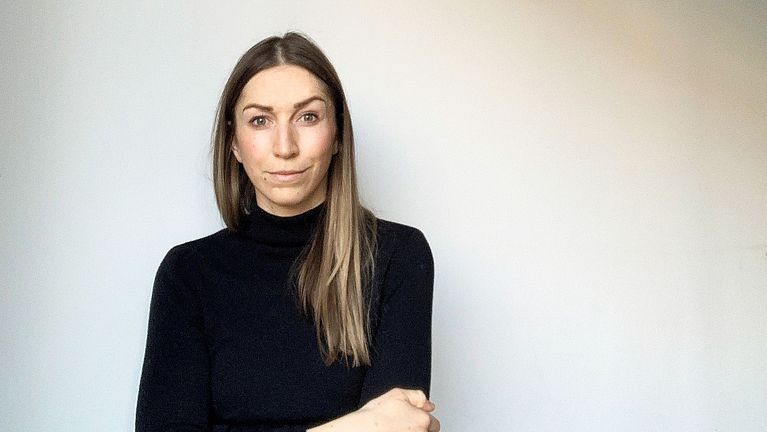 A woman with long brown hair, wearing a black turtleneck, stands with her arms crossed against a plain background.