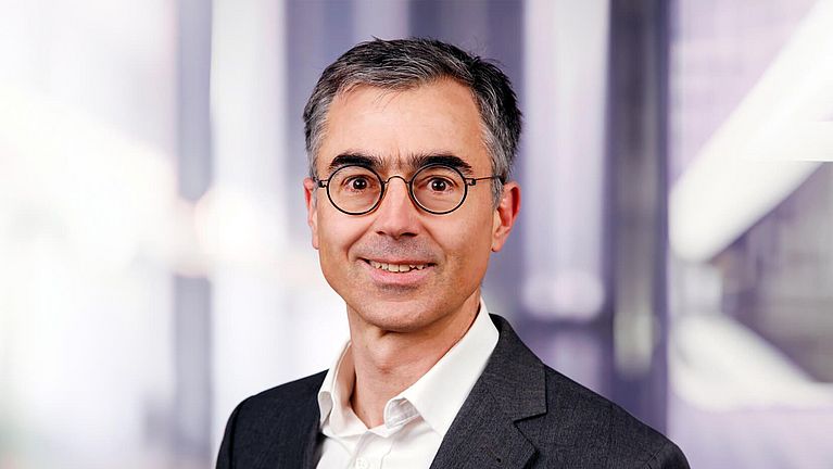 Man with short gray hair and glasses, wearing a dark suit and white shirt, smiles against a blurred background.